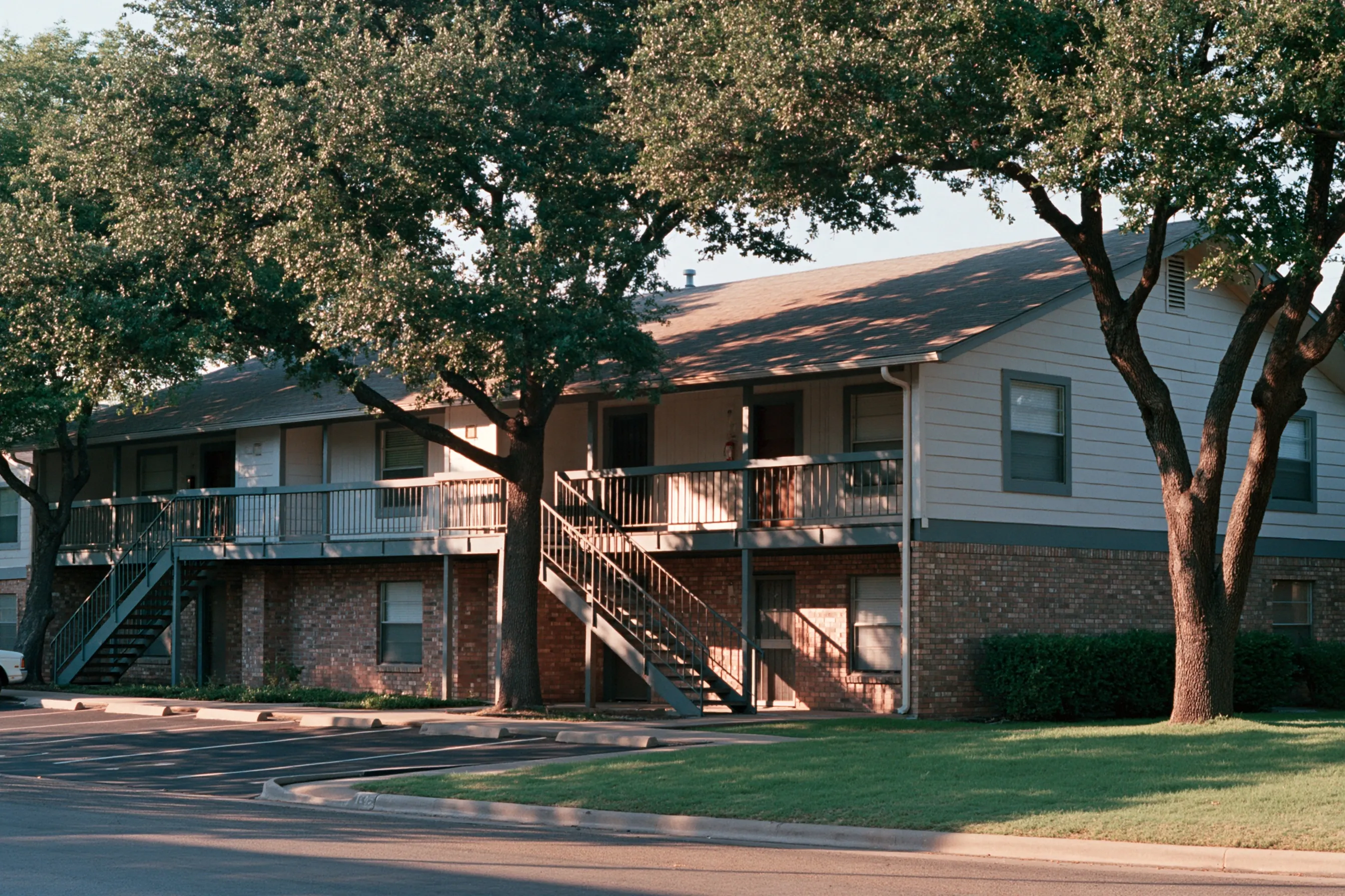 Two-story 1980s garden apartment building with exterior walkways, wood siding and brick, North Texas, live oaks, no people
